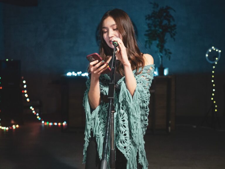 A young woman singing into a microphone on stage while looking at her phone