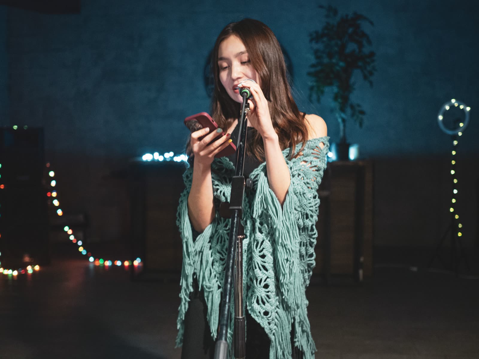 A young woman singing into a microphone on stage while looking at her phone
